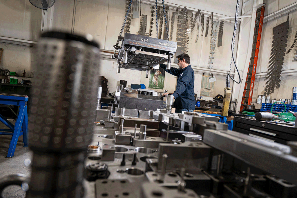A side view of a young man wearing goggles operating a stamping press beyond out of focus die tools.