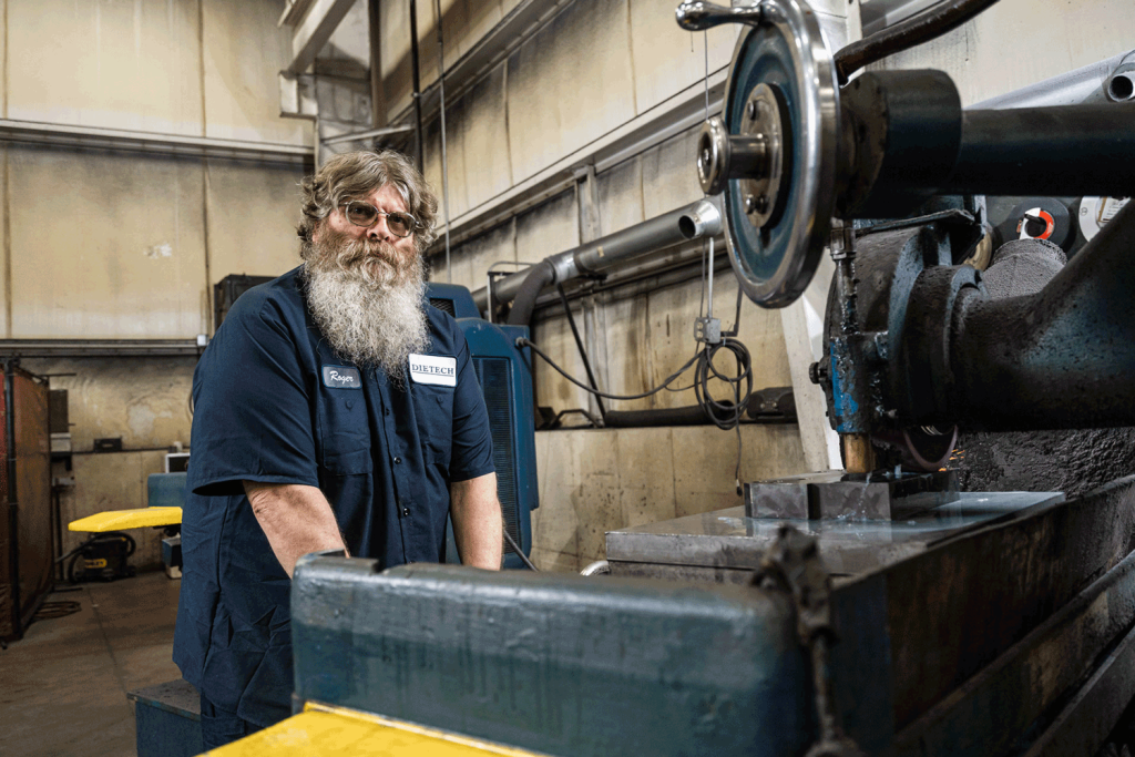 A bearded man wearing glasses stands before a machine, exuding confidence and expertise.